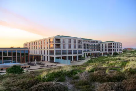 Außenansicht des modernen Hotelgebäudes des A-ROSA Sylt bei Sonnenuntergang. Das Hotel A-ROSA Sylt ist umgeben von Dünenlandschaften und bietet einen Außenpoolbereich mit Liegestühlen. Die großflächigen Fenster und Balkone des Hotels spiegeln das warme Abendlicht wider, während der Himmel in sanften Rosa- und Blautönen getaucht ist, was eine ruhige und luxuriöse Atmosphäre erzeugt.