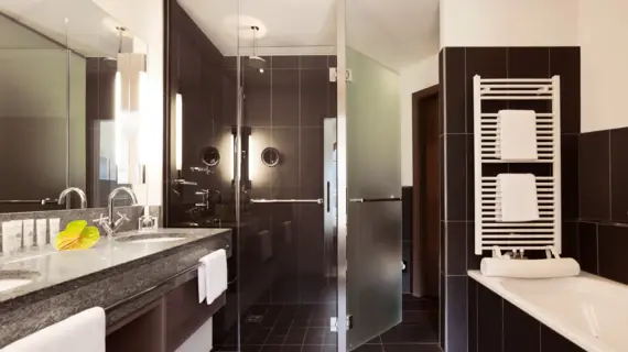A bathroom with dark tiles, a large shower, a bathtub and a washbasin. A towel warmer with two white towels hangs above the bathtub.