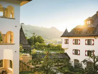 Ein Schlosshotel von außen mit Türmen und Balkonen. Im Hintergrund geht die Sonne unter und es ist eine Berglandschaft zu sehen.