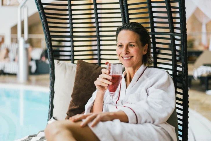 Eine lächelnde Frau, eingehüllt in einen weißen Bademantel, entspannt sich in einem großen nestförmigen Rattansessel und hält ein Glas mit einem roten Getränk, im Hintergrund ein Innenpoolbereich.