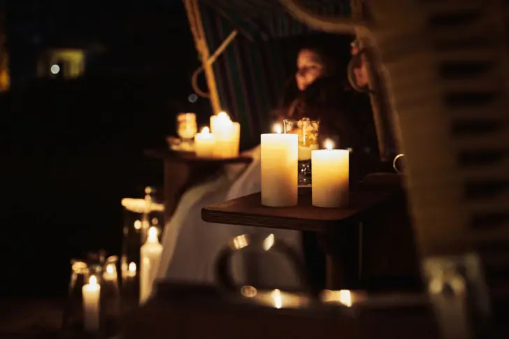 Ein stimmungsvolles Kerzenlicht-Setting in der Nacht: Mehrere brennende Kerzen stehen auf einem kleinen Holztisch neben einem Strandkorb. Im Hintergrund sitzt eine Person in eine Decke gehüllt, während weitere Kerzen in Gläsern für eine warme, gemütliche Atmosphäre sorgen.