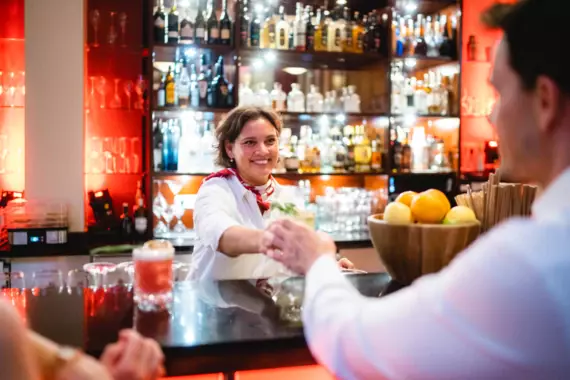 Eine freundlich lächelnde Barkeeperin reicht einem Gast an der Bar ein Getränk. Im Hintergrund ist eine gut sortierte Bar mit einer Vielfalt an Flaschen und einem warmen Licht, das die Szene beleuchtet. Im Vordergrund ist der Rücken eines Gastes zu sehen, der am Tresen sitzt.