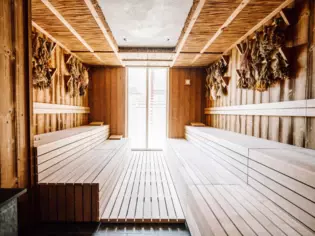 Das Bild zeigt das Innere einer Sauna, die traditionell mit Holz verkleidet ist. Die Wände sind aus naturbelassenem Holz gefertigt, welches eine warme, gemütliche Atmosphäre schafft. An der Decke ist eine besondere Verkleidung aus Reet zu erkennen, die eine rustikale Note hinzufügt. Die mehrstufigen Saunabänke sind hell und laden zum Verweilen ein. An der Wand hängen verschiedene Bündel aus getrockneten Kräutern, die sowohl als Dekoration dienen als auch ein authentisches Saunaerlebnis durch ihren Duft unterstützen. Ein Fenster lässt natürliches Licht herein und unterstreicht die warme und einladende Atmosphäre des Raumes.