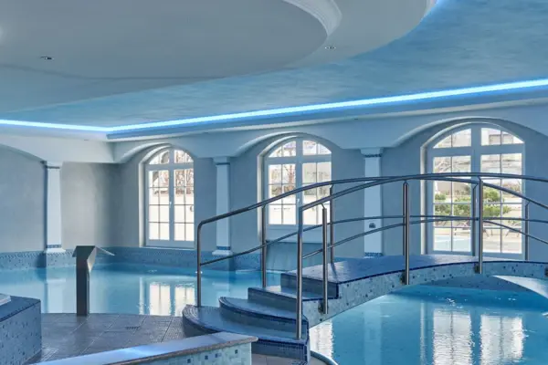 An indoor pool in light blue tones with lots of large windows and a small bridge leading over the pool. 