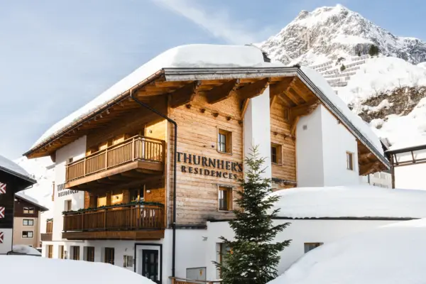 Außenansicht der verschneiten Thurnher’s Residences in Zürs am Arlberg vor alpiner Bergkulisse bei Sonnenschein – ein stilvoller Rückzugsort für den Winterurlaub.