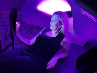 Eine Person liegt halb aufgerichtet auf einer Hydrojetliege in einem Wellnessbereich, beleuchtet von violettem Licht, und interagiert mit einem Steuergerät oder Display.