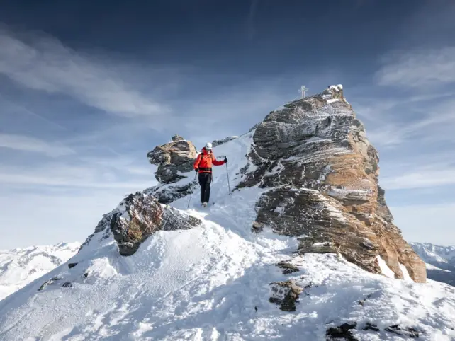 Ein Mensch in roter Jacke und Skiausrüstung geht eine Schneebedeckten Gipfel hinunter. 