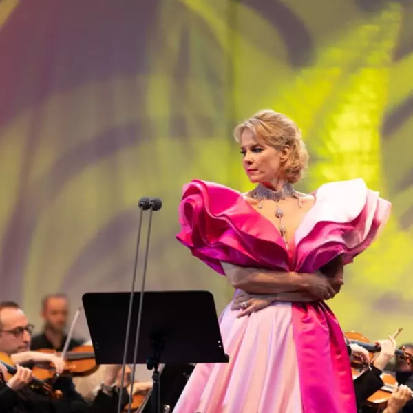 Eine Sopranistin in einem auffälligen pinken Kleid, umgeben von einem Orchester und einem farbenfrohen Hintergrund. 