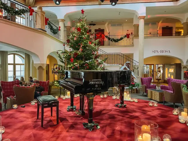 Eine weihnachtliche Innenansicht der Lobby. Auf einem roten Teppich steht ein brauner Flügel und ein Tannenbaum. Drumherum stehen Sessel, um es sich gemütlich zu machen. 