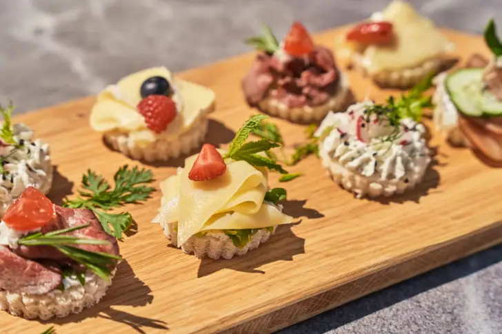 Ein Holzbrett mit verschiedenen Speisen, darunter Fingerfood, Früchte und Käse, kunstvoll garniert auf einem Tisch.