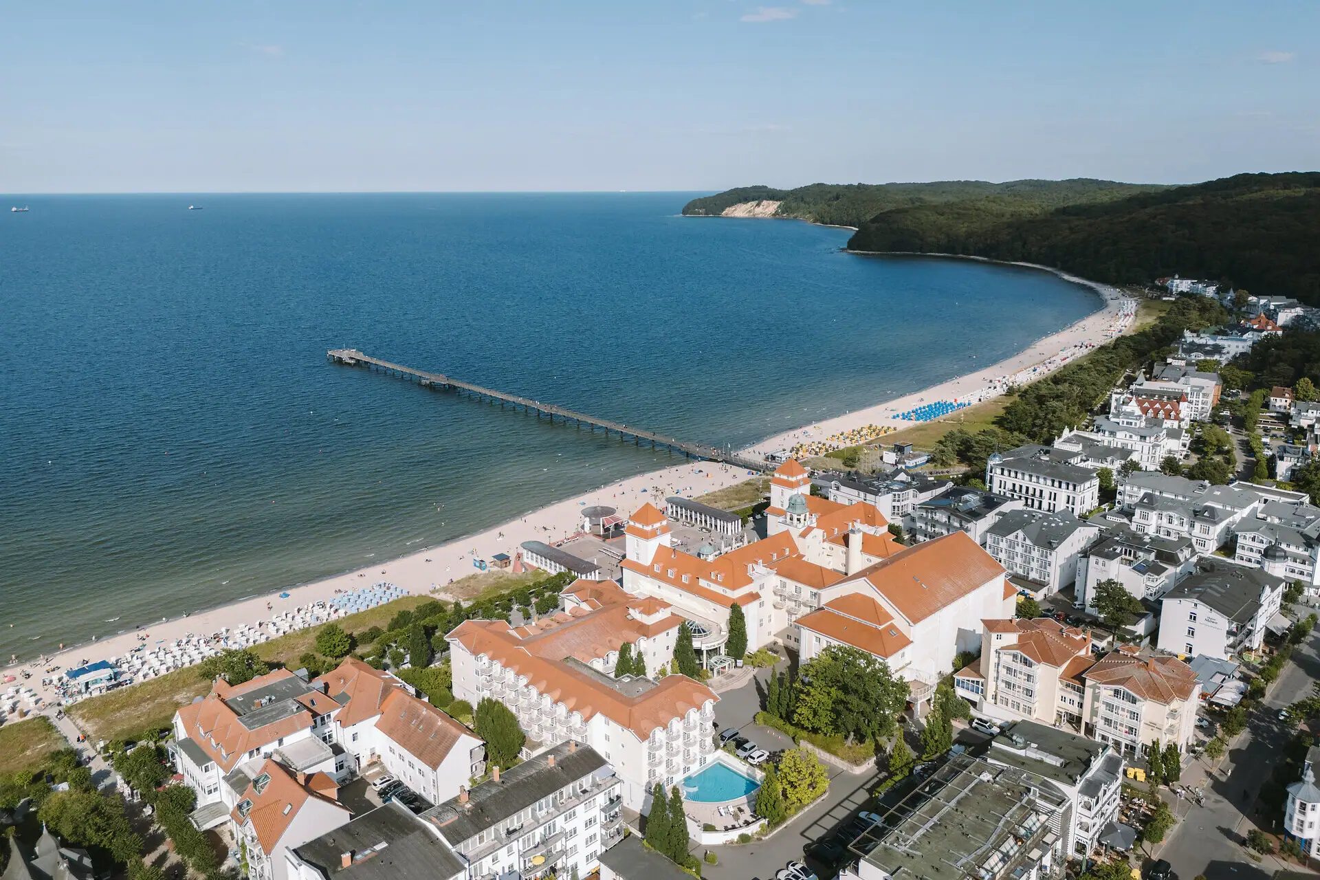 Die Küste von Binz aus der Luft fotografiert mit einem Strand, Gebäuden und einem Pier bei blauem Himmel.