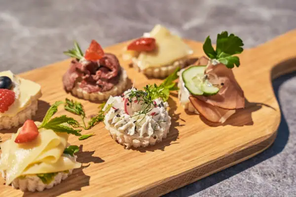Fein garnierte Canapés auf einem Holzbrett, belegt mit Käse, Roastbeef, Frischkäse, Schinken und frischen Kräutern, angerichteten als Fingerfood bei Sonnenschein. 