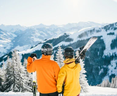 Zwei Personen, eine mit einer orangenen und eine mit einer gelben Skijacke stehen in einem Skigebiet auf einem Berg und schauen in die Ferne. Beide Personen haben einen Skihelm auf und tragen ihre Skier auf der Schulter.