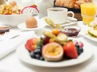 Ein heller, reichgedeckter Frühstückstisch, auf dem sich Kaffee, Orangensaft, ein Ei und ein Teller voller Früchte befindet. 