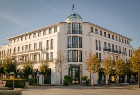 Elegantes weißes Hotelgebäude mit moderner Architektur und einem runden Glasdach, umgeben von herbstlichen Bäumen und einer gepflegten Umgebung, unter klarem blauem Himmel.