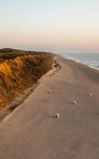 Eine Luftaufnahme von einem breiten Sandstrand mit Klippe und Wasser im Hintergrund bei Sonnenuntergang.