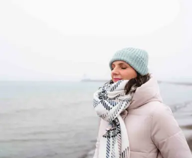 Eine Frau steht in warmer Winterkleidung mit geschlossenen Augen an einem nebeligen Strand und atmet tief durch. 