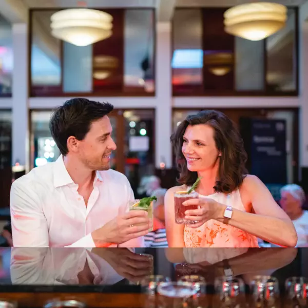 Ein Paar genießt gemeinsam einen entspannten Abend in einer Bar. Sie sitzen an der Bartheke und halten jeweils einen Cocktail in der Hand. Der Mann, gekleidet in ein weißes Hemd, lächelt der Frau gegenüber, die ein sommerliches, apricotfarbenes Top trägt und ihm fröhlich zulächelt. 