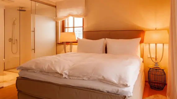 Ein Schlafzimmer mit direktem Zugang zum Bad. In der Mitte des Zimmers steht ein großes Polsterbett mit weißer Bettwäsche und links und rechts davon stehen jeweils ein Nachttisch mit großen Lampen.