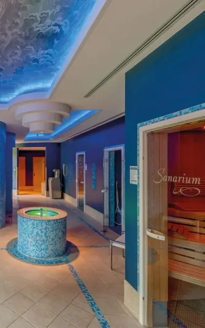 Innenansicht eines stilvoll gestalteten Spa-Bereichs mit blauer Deckenbeleuchtung, einem runden Wasserbecken in der Mitte, einer Sanarium-Sauna auf der rechten Seite.
