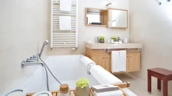 A bright bathroom with a bathtub, a large washbasin with two washbasins and a small stool. A towel warmer can be seen in the background.