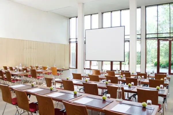 Ein Tagungsraum mit Tischen und Stühlen vor einer großen Glasfront, wodurch viel Tageslicht in den Raum kommt.