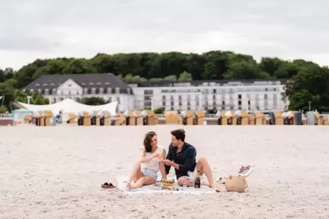 Ein junges Paar genießt ein romantisches Picknick am Strand. Sie sitzen auf einer Decke im Sand, umgeben von einem Picknickkorb und verschiedenen Leckereien. Im Hintergrund ist das A-ROSA Hotel in Travemünde zu sehen, hinter einer Reihe bunter Strandkörbe. Die Szene strahlt eine entspannte und glückliche Atmosphäre aus, während das Paar in vertrauter Zweisamkeit ein Gespräch führt und die gemeinsame Zeit schätzt.