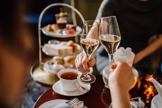 Zwei Personen stoßen mit Sektgläsern an, vor ihnen steht ein stilvoll arrangierter Afternoon-Tea mit einer Etagere voller Leckereien, einer Teetasse und einem Teekännchen.