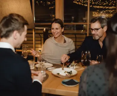 Eine Gruppe aus zwei Paaren sitzt bei gemütlichem Licht zusammen um einen Tisch herum, hält Weingläser in ihren Händen und teilt sich eine Nachspeise. 