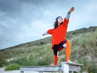 Tina Molsen steht mit warmen Yogasachen auf einem Holzpodest am Strand mit den Dünen im Hintergrund und macht eine Übung. 