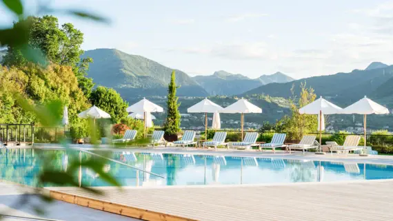 A pool surrounded by sun loungers and parasols, flanked by trees under a clear sky.