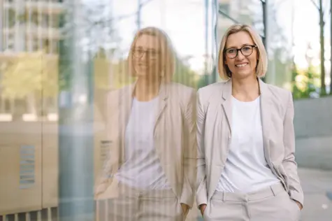 Eine Frau in hellem Anzug lehnt Lächeln an einer Glasscheibe. 