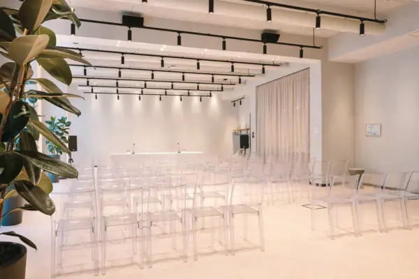 Ein Tagungsraum mit transparenten Stühlen und einem Pult vorne. Das Pult ist mit Mikrofonen ausgestattet. Außerdem stehen am Rand jeweils Lautsprecher.