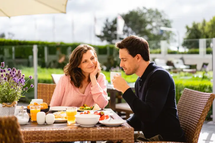 Zwei Personen genießen ein Frühstück im Freien; die Frau lächelt, während sie den Mann beobachtet, der gerade von seiner Tasse trinkt, umgeben von einer idyllischen Umgebung mit frischen Früchten und Säften auf dem Tisch.