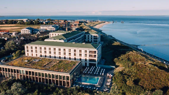 Luftaufnahme eines modernen Resortkomplexes mit begrüntem Dach am Meer. Das Hotel auf Sylt ist von üppigen Grünflächen und einem Sandstrand flankiert. Der Blick erstreckt sich bis zum Horizont über die ruhige See. Die angrenzende Ortschaft fügt sich harmonisch in die Küstenlandschaft ein.
