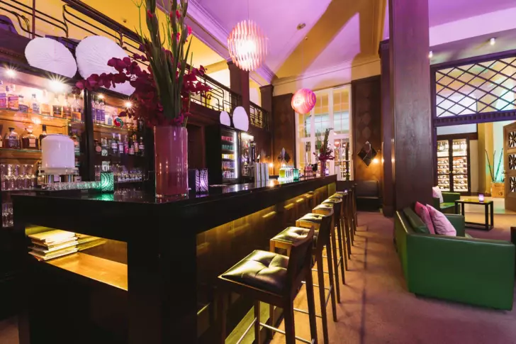 Stimmungsvolle Bar mit dunklen Holzmöbeln, leuchtenden Orchideen und Papierlaternen. Eine Reihe von Barhockern steht vor der Theke, die mit grünen Lichtakzenten beleuchtet ist, während im Hintergrund eine gemütliche Lounge-Ecke mit grünen Sofas und einem beleuchteten Weinregal zu sehen ist.