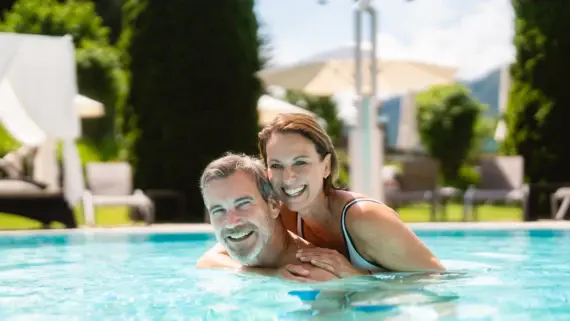 Ein lächelndes Paar umarmt sich im Pool, umgeben von einer grünen Gartenlandschaft und Liegestühlen im Hintergrund bei sonnigem Wetter