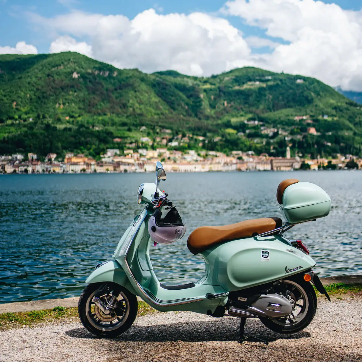 Eine mintgrüne Vespa steht am Ufer des Gardasees, im Hintergrund die malerische Uferstadt und die grünen Hügel der Region.