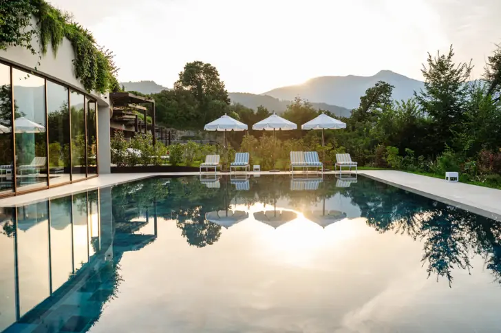 Swimmingpool im Freien umgeben von Stühlen und Sonnenschirmen mit Bäumen und einem klaren Himmel mit untergehender Sonne im Hintergrund.