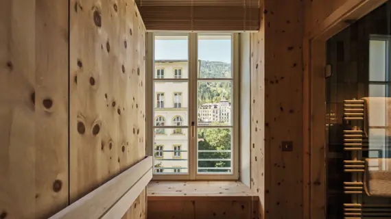 Eine Sauna mit einem Fenster, durch das ein anderes helles Haus sowie eine Berglandschaft mit vielen Tannen zu sehen ist.