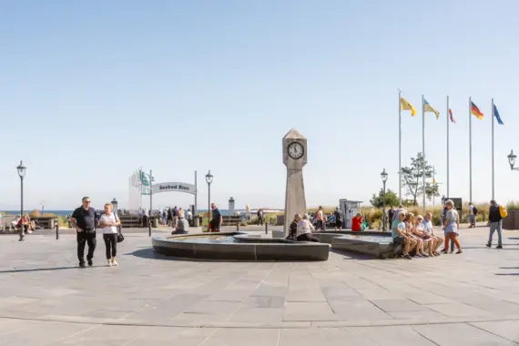 Eine Gruppe von Menschen auf einem Platz mit einem Uhrturm und einem Brunnen.