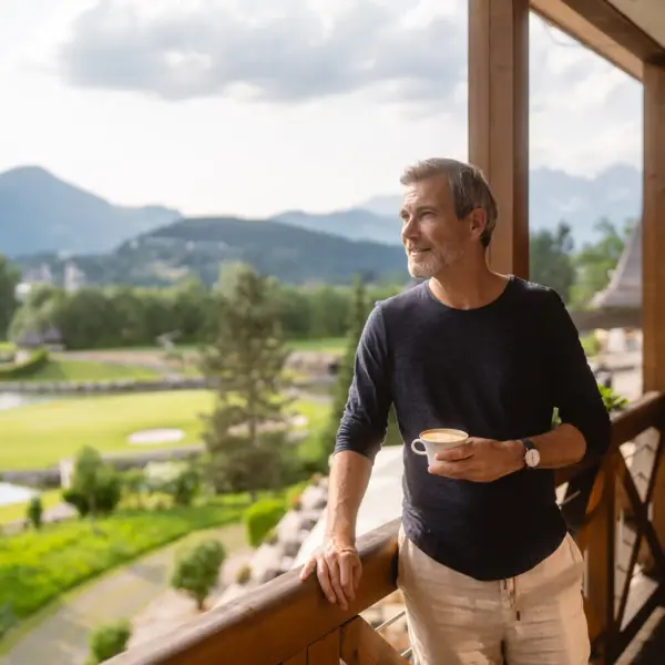 Mann steht mit einem Kaffee auf dem Balkon und schaut in die Berge. 
