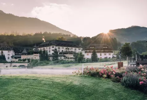 Morgendämmerung im A-ROSA Hotel in Kitzbühel mit aufgehender Sonne, die hinter den malerischen Bergen hervorleuchtet. Ein leichter Nebel schwebt über dem ruhigen See vor dem traditionell alpenländischen Hotelgebäude, umgeben von einem üppigen Garten mit einer Vielfalt an Blumen in voller Blüte.