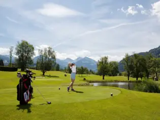 Zwei Personen auf einem gepflegten Golfplatz mit Blick auf eine beeindruckende Alpenkulisse, umgeben von grünen Bäumen und einer idyllischen Landschaft unter einem teilweise bewölkten Himmel.