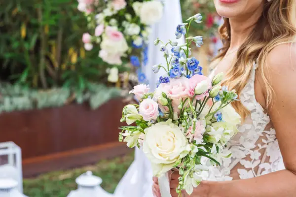 Eine Braut im Spitzenkleid hält einen farbenfrohen Hochzeitsstrauß mit Rosen, Lisianthus und blauen Akzenten.