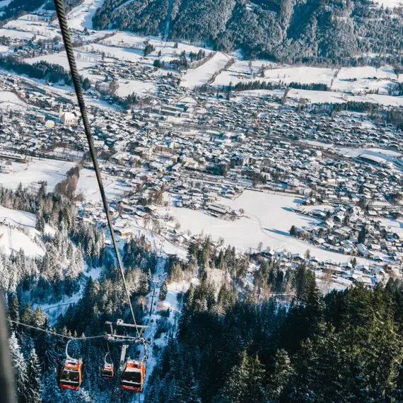 Eine Seilbahngondel, umgeben von einer verschneiten Berglandschaft, schwebt über einem malerischen Dorf. Der Blick aus der Perspektive der Seilbahn zeigt den verschneiten Ort Kitzbühel mit seinen dicht gedrängten Häusern und Straßen, eingebettet in die winterliche Alpenlandschaft. Im Hintergrund erhebt sich ein markanter Berg, dessen Gipfel im Sonnenlicht glänzt.