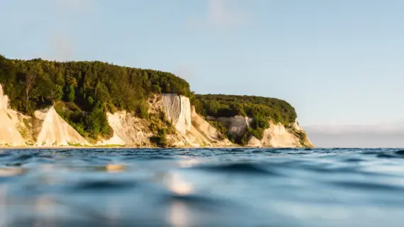 The bright chalk cliffs are covered in green vegetation and protrude from the bright blue water. 