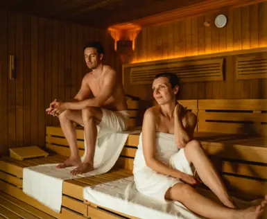  Das Bild zeigt ein Paar, das in einer Sauna entspannt. Die Sauna ist traditionell gestaltet mit Holzverkleidung und sanftem Licht, das eine ruhige und warme Atmosphäre schafft. Beide Personen sitzen ruhig, umgeben von der Wärme des Raumes, was typisch für das Saunaerlebnis ist, das sowohl Entspannung als auch Wohlbefinden fördert. Solche Momente sind ideal, um vom stressigen Alltag abzuschalten und die Gesundheit zu pflegen.