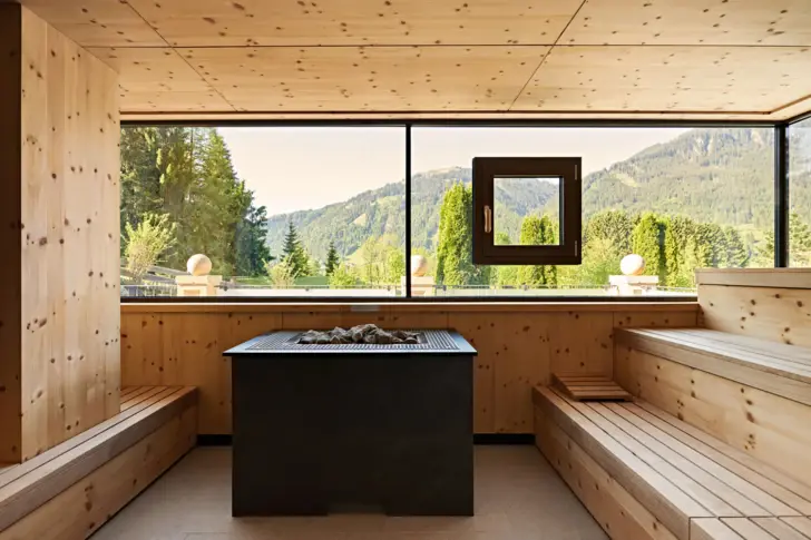 Eine moderne Sauna mit großen Fenstern, die einen Blick auf grüne Berge bieten. Die Wände und Bänke sind aus hellem Holz.  Im Zentrum steht ein schwarzer Saunaofen mit Steinen.