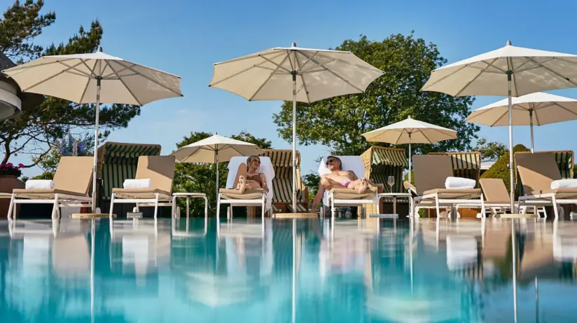 Ein Mann und eine Frau liegen auf Liegestühlen an einem Pool unter einem Sonnenschirm, umgeben von Bäumen.